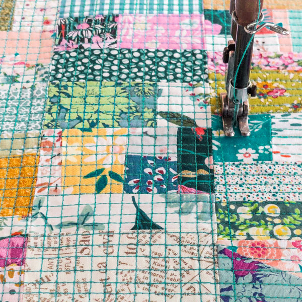 Detalhe do processo de quilting sobre uma composi&ccedil;&atilde;o de retalhos de tecidos coloridos e estampados, com costuras em grelha que criam textura e unem os diferentes fragmentos.