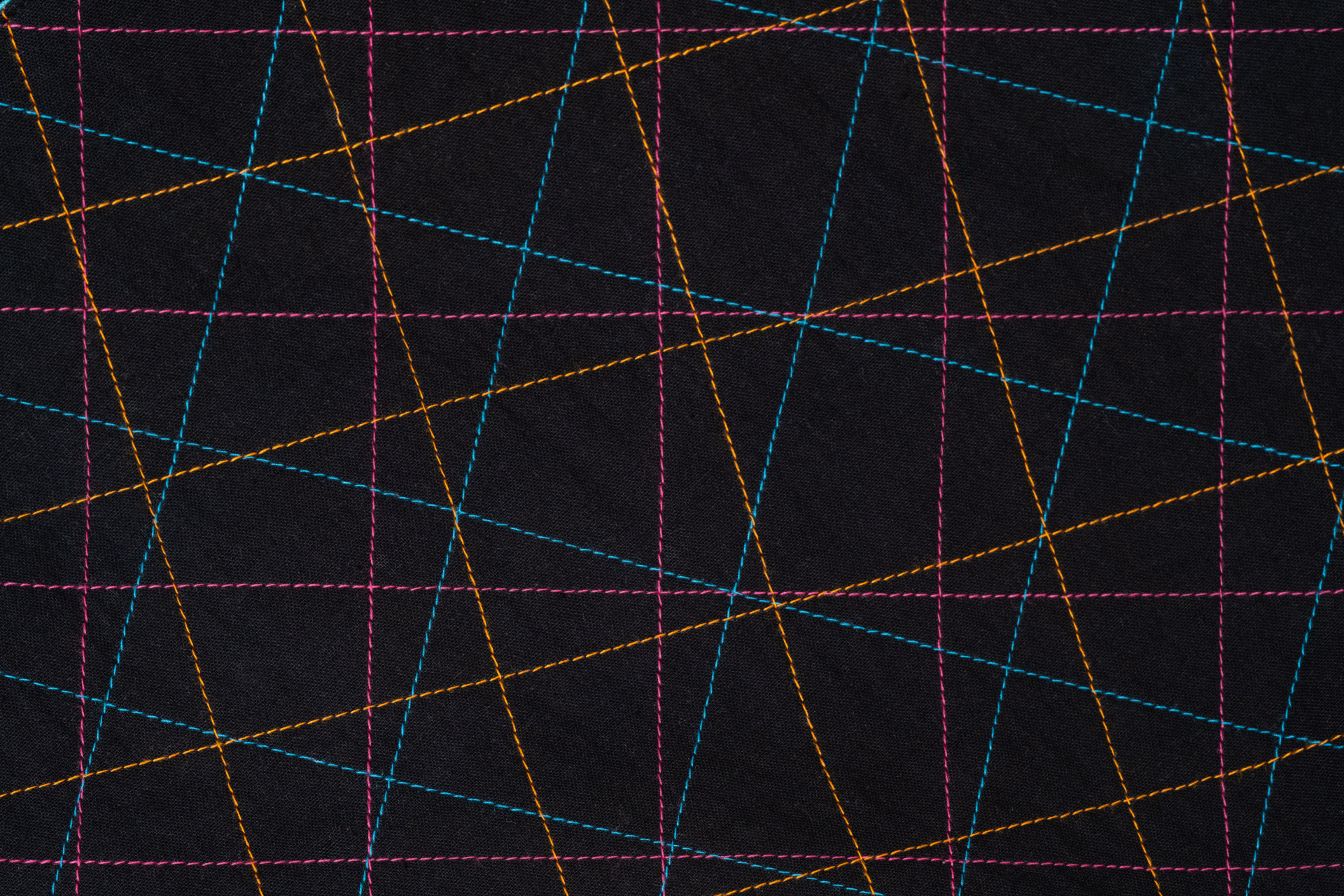 Detalhe de quilting geométrico sobre tecido preto, com linhas de costura em rosa, azul e laranja que se cruzam em diferentes ângulos, criando uma composição gráfica.