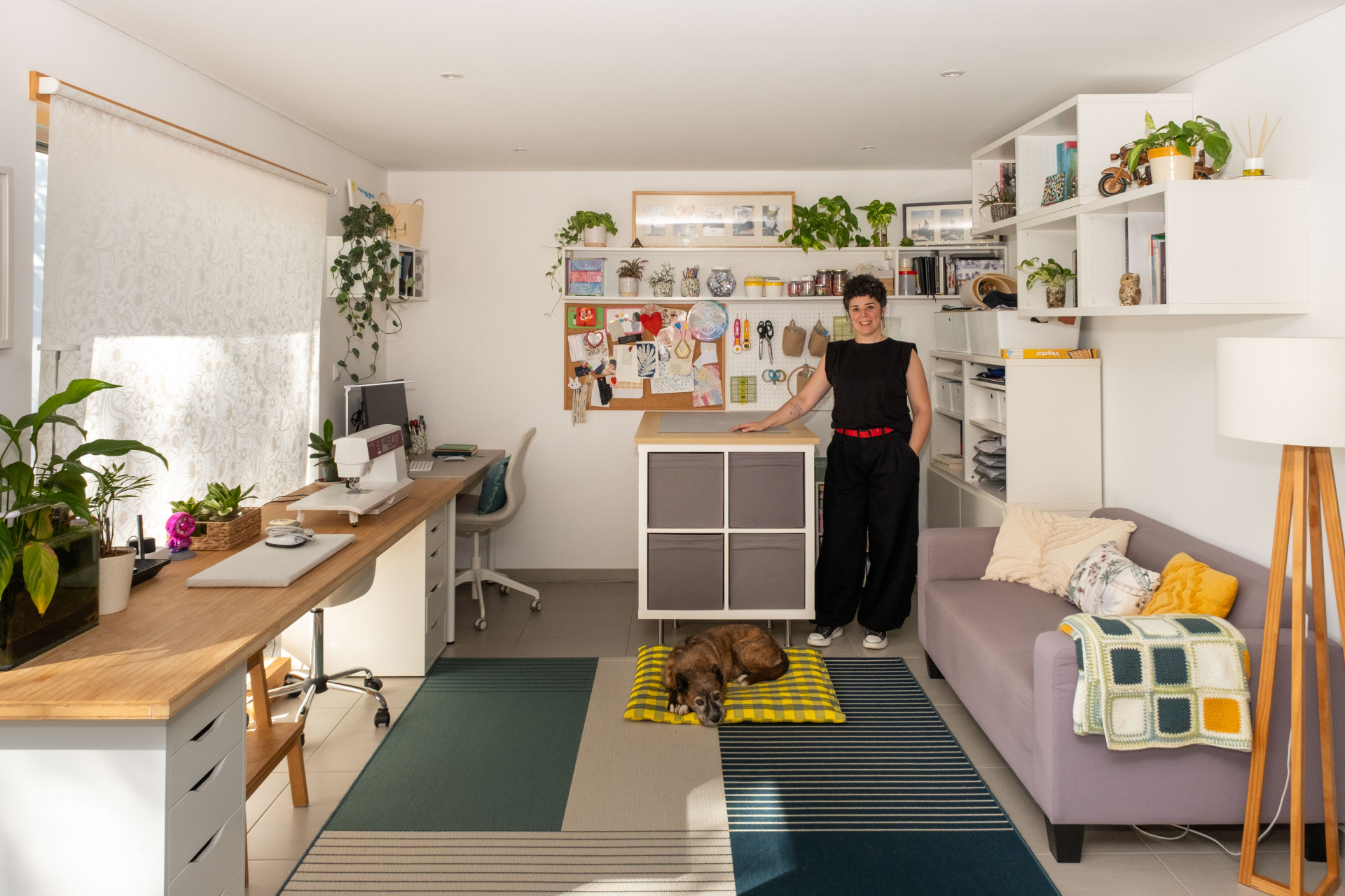 Vista geral do atelier Pontos Studio, com Isabel Moreira junto à área de trabalho, máquinas de costura, materiais criativos, plantas e um cão a descansar no centro do espaço.